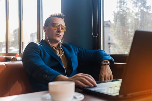 A business serious man businessman a stylish of Caucasian appearance, in sunglasses, works in a laptop or computer, sitting at a table by the window in a cafe and listening to music on headphones.