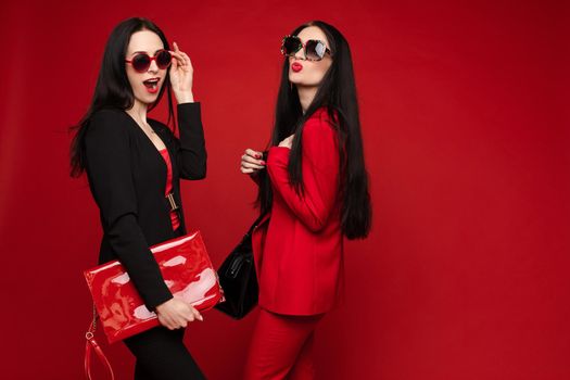 Side view of two brunettes wearing black and red smart suits and glasses standing back to back on isolated background. Stylish women posing and looking at camera in studio. Concept of glamour.