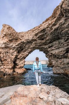 A woman in a blue jacket stands on a rock above a cliff above the sea and looks at the raging ocean. Girl traveler rests, thinks, dreams, enjoys nature. Peace and calm landscape, windy weather