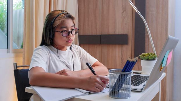  Asian girl in headphones sit at desk study online on laptop. Kid wear headset handwriting in notebook learning using internet lessons on quarantine. Girl student learning virtual internet online class from school due to covid pandemic.
