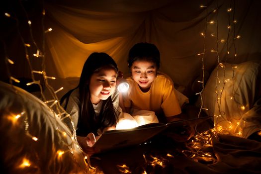 Family concept. elder sister and sister reading book with flashlight together before bedtime. Sister read story book together in bed sheet tent. focus selective little sister. With film grain effect