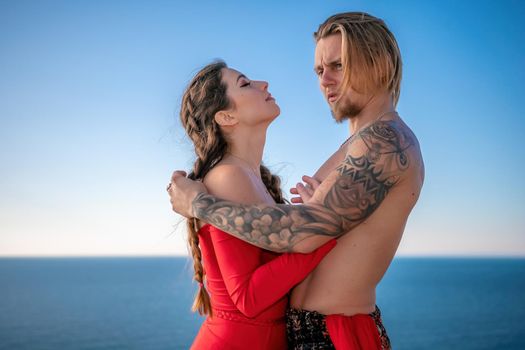 Portrait of tattooed man and sensual young brunette woman in red dress, happily dancing outdoors isolated on blurry sea background with natural bokeh in soft warm sunset backlight. Selective focus