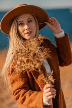 A woman walking along the coast near the sea. An elegant lady in a brown coat and a hat with fashionable makeup walks on the seashore.