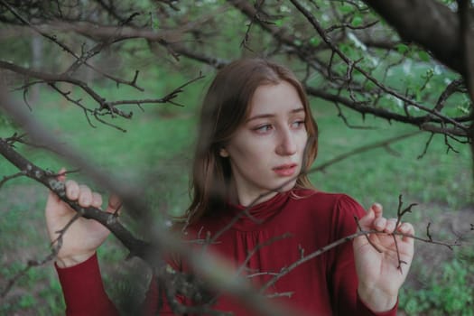 a blonde girl in red looks into the camera through the branches of a tree