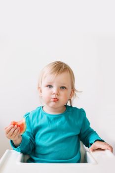 portrait of a funny little baby girl sitting in a high chair and eating watermelon.