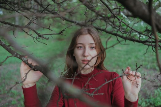 a blonde girl in red looks into the camera through the branches of a tree