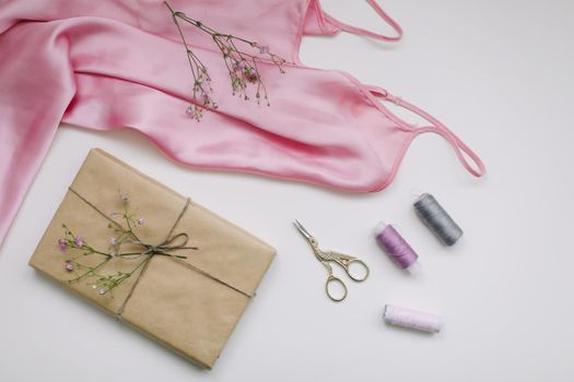 Composition with pink silk fabric, gift in craft paper and sewing tools, threads and scissors on white background. Flat lay, top view. Hobby, leisure concept.