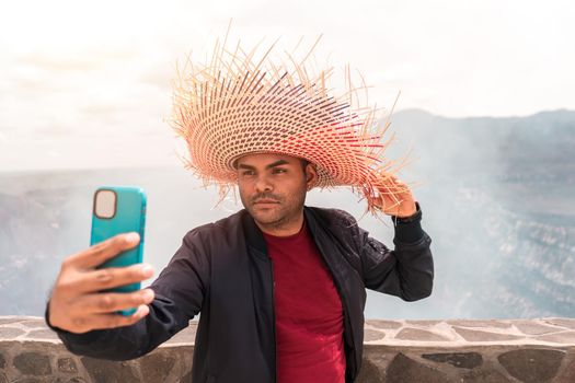 Mestizo latin man taking a selfie with his smartphone wearing a colorful hat in front of the crater of the Masaya volcano in Nicaragua, an important tourist destination in Latin America.
