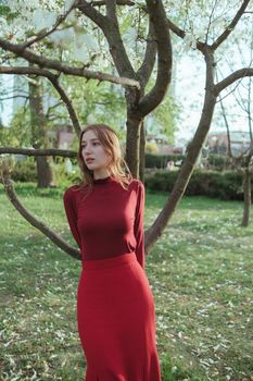 A blonde girl in red leans on a tree in the park. the concept of unity with nature