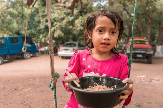 Latin girl sitting on a swing eating from a plastic bowl. Concept of childhood, nutrition and food in rural areas of Nicaragua Latin America