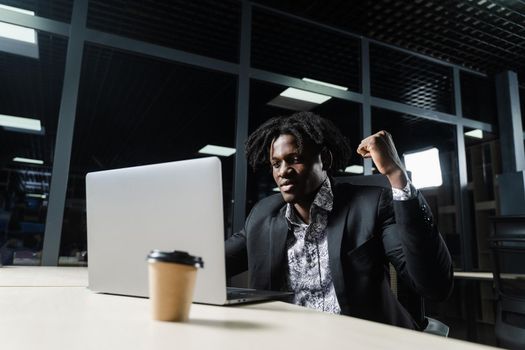 Happy black man with clenched fists is looking on the screen of laptop and rejoice finishing his job