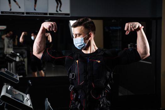 Man in EMS suit and medical mask in gym. Protection from coronavirus covid-19. Sport training in electrical muscle stimulation suit at quarantine period