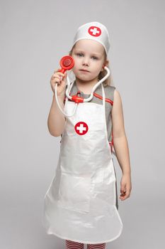 Confident little girl posing in doctor uniform coat holding stethoscope enjoying playing. Cute baby child wearing nurse clothing and hat looking at camera at white studio background