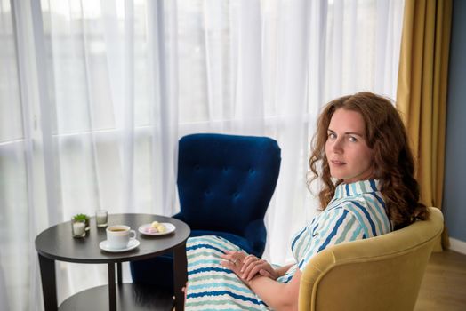 Young woman at home sitting on modern chair in front of window relaxing in her living room and drinking coffee or tea with sweets or desert. Relax before new business day.