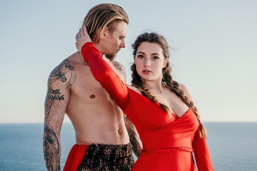 Portrait of tattooed man and sensual young brunette woman in red dress, happily dancing outdoors isolated on blurry sea background with natural bokeh in soft warm sunset backlight. Selective focus