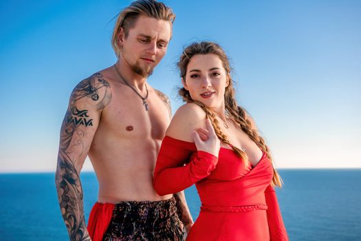 Portrait of tattooed man and sensual young brunette woman in red dress, happily dancing outdoors isolated on blurry sea background with natural bokeh in soft warm sunset backlight. Selective focus