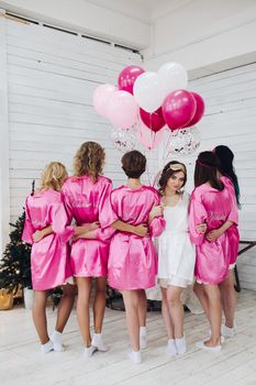 Studio shot of bride-to-be in white robe standing with her bridesmaids in pink robe back to camera with bunch of air balloons.