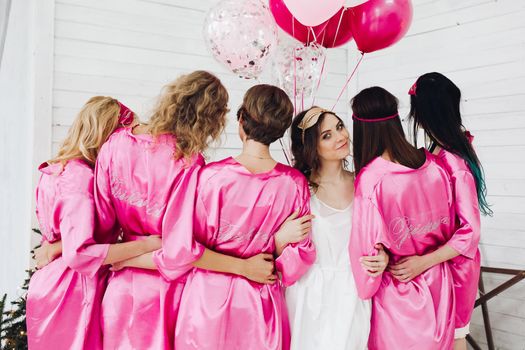 Studio shot of bride-to-be in white robe standing with her bridesmaids in pink robe back to camera with bunch of air balloons.