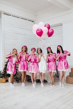 Full length portrait of happy lovely girlfriends in pink robes and sleeping masks jumping and having fun at hen party. Bride-to-be in white robe holding pink air balloons with smile standing on tiptoes.