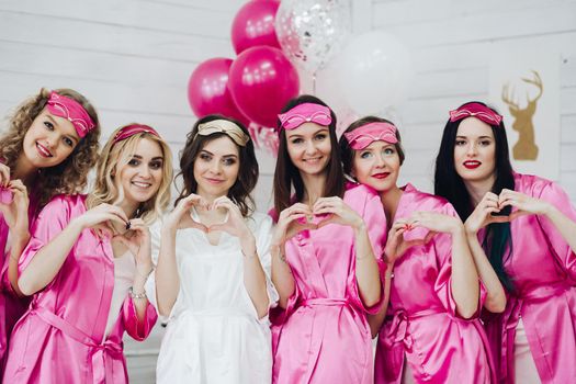 Portrait of group of happy girlfriends in bright pink robes and sleep masks showing hearts with hands at celebration of bridal shower.