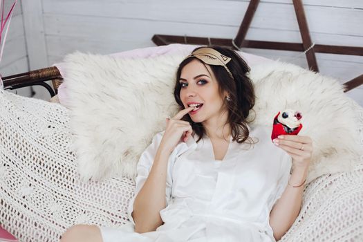 Portrait of brunette bride-to-be in white silk bathrobe and cat sleeping mask biting her finger with cupcake in other hand sitting in cozy armchair with fur cover..