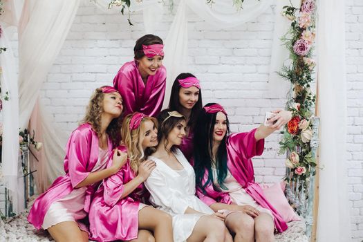 Group of lovely girls in pink robes and sleeping masks taking selfie with beautiful bride-to-be in white robe.