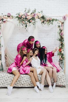 Group of lovely girls in pink robes and sleeping masks taking selfie with beautiful bride-to-be in white robe.