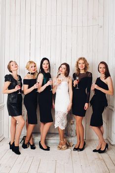 Portrait of beautiful elegant bridesmaids in black dresses toasting glasses of champagne with bride-to-be in lovely white dress.