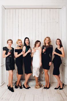 Portrait of beautiful elegant bridesmaids in black dresses toasting glasses of champagne with bride-to-be in lovely white dress.