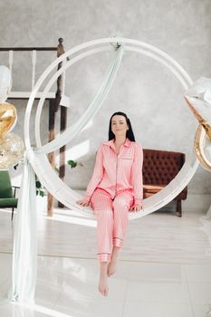 Studio portrait of beautiful young black-haired girl in pink striped pyjamas sitting elegantly on hanging roung chair surrounded by white air balloons.