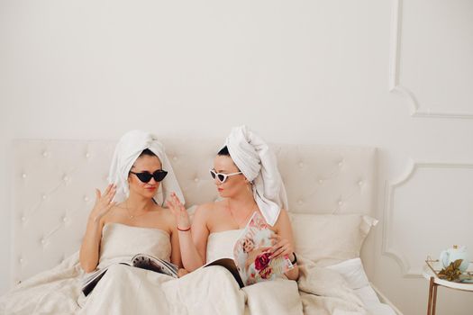 Stock photo of incognito women in bath towels on head and sunglasses holding fashion magazines sitting in bed. Fashion victims in the morning. Fashion divas preparing for the day reading fashion journals in bed.