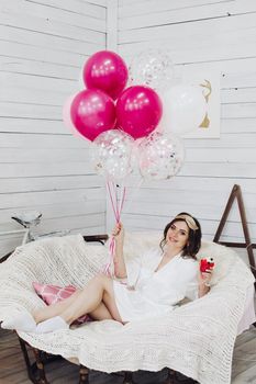 Portrait of attractive bride-to-be in white silk robe with sleeping mask on head holding delicious berry cake while relaxing in cozy white armchair.