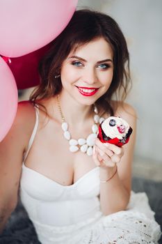 Portrait of beautiful happy bride-to-be with make up in white dress holding berry muffin and air balloons. Bridal shower.