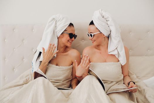 Two happy glamour woman friend wearing towel relaxing together. Fashion smiling girl in sunglasses sitting on bed in bedroom holding magazine enjoying having good time indoor medium shot