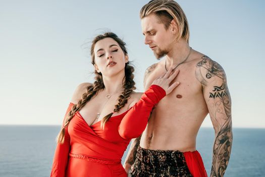 Portrait of tattooed man and sensual young brunette woman in red dress, happily dancing outdoors isolated on blurry sea background with natural bokeh in soft warm sunset backlight. Selective focus