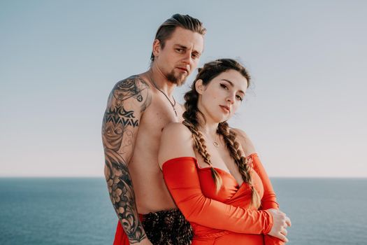 Portrait of tattooed man and sensual young brunette woman in red dress, happily dancing outdoors isolated on blurry sea background with natural bokeh in soft warm sunset backlight. Selective focus