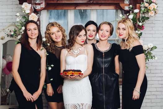 Portrait of beautiful bridesmaids in black dresses with attractive bride-to-be in the middle holding delicious berry cake. Background is decorated with flowers.