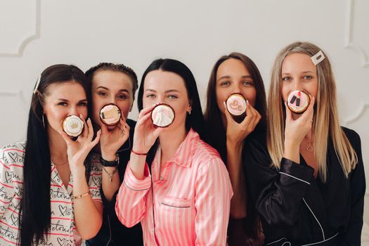 Stock photo of pretty bridesmaids in pyjamas holding delicious handmade cupcakes in front of their lips looking at camera. Celebrating hen party together. Best friends.