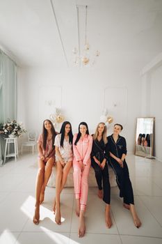 Stock photo of five girls in pajamas eating sweet lollipops sitting close on big white bed in beautiful white room with inflated air balloons in shape of wedding ring, hearts and stars.