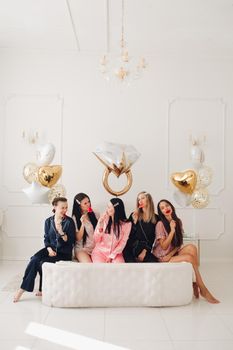 Stock photo of five girls in pajamas eating sweet lollipops sitting close on big white bed in beautiful white room with inflated air balloons in shape of wedding ring, hearts and stars.