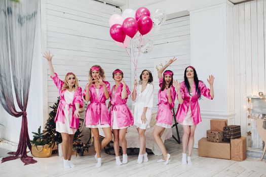 Full length portrait of happy lovely girlfriends in pink robes and sleeping masks jumping and having fun at hen party. Bride-to-be in white robe holding pink air balloons with smile standing on tiptoes.