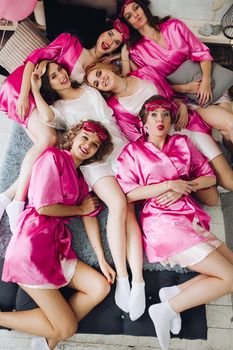 Overhead of pretty young ladies in pink bathrobes lying on bed with bride-to-be in white dress. Girlfriends showing tongues out and smiling. Bachelorette party.