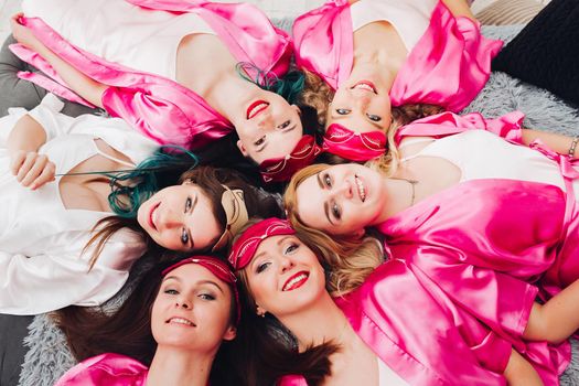 Overhead of happy pretty bridesmaids in pink robes with bride to be and sleeping masks on bed celebrating bridal showers.