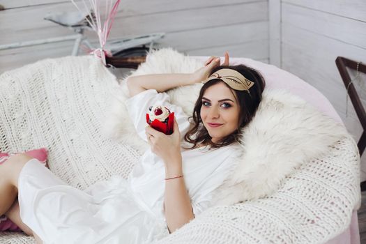 Portrait of attractive bride-to-be in white silk robe with sleeping mask on head holding delicious berry cake while relaxing in cozy white armchair.
