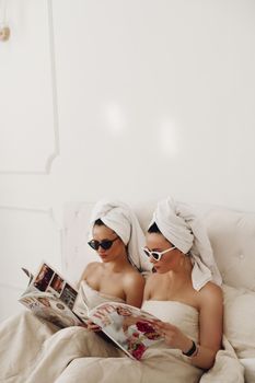 Young females in sunglasses and towels sitting on bed while reading magazines. Care, leisure and relaxation time