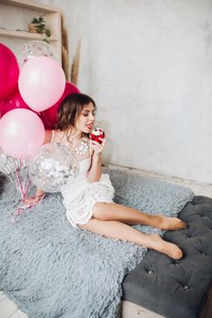 Portrait of pretty young bride-to-be in white dress sitting on sofa with muffin and pink air balloons. Hen party.