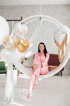 Studio portrait of beautiful young black-haired girl in pink striped pyjamas sitting elegantly on hanging roung chair surrounded by white air balloons.