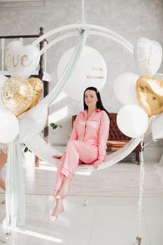 Studio portrait of beautiful young black-haired girl in pink striped pyjamas sitting elegantly on hanging roung chair surrounded by white air balloons.