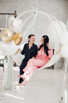 Two happy female friend in pajamas sitting on stylish armchair at luxury interior. Smiling girlfriends enjoying friendship surrounded by festive party event having positive emotion