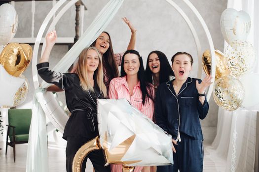 Five friends celebrating the party dedicated to a bachelorette in the middle. Hen party celebration. Girls laughing and smiling with black-haired girl in the middle holding a big balloon ring.